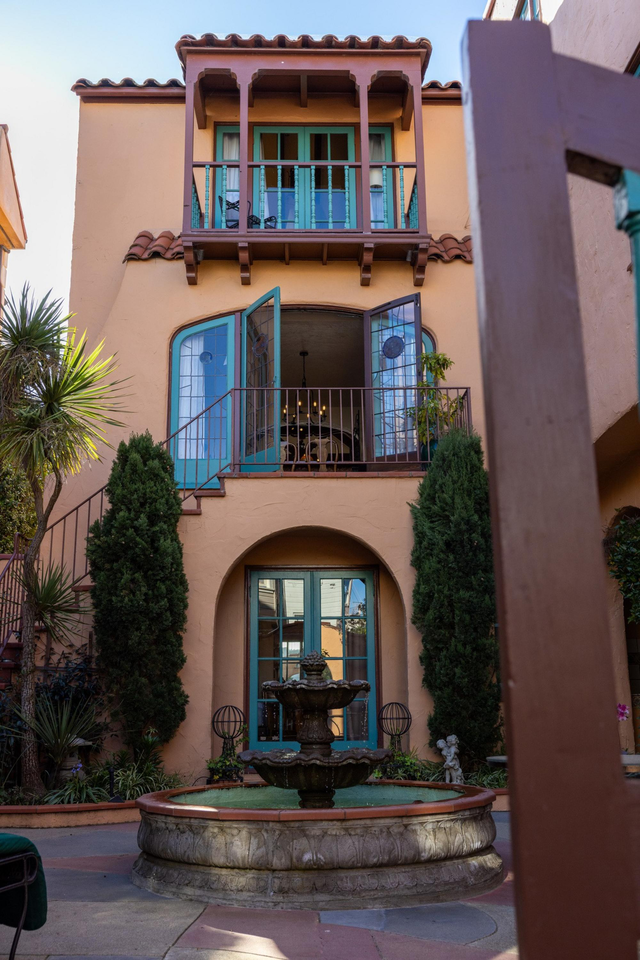 A two-story building with a balcony and open teal doors overviews a courtyard with a central stone water fountain surrounded by tall green plants.